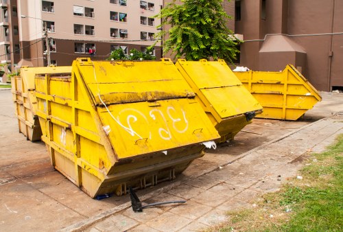 A large yellow metal skip with a weathered surface, placed on a paved area adjacent to a multi-storey residential building. The skip displays visible rust marks and faded white graffiti on its side. It is positioned with the open top facing upward, and the rear end extends outward, indicating recent use for waste collection. The environment includes a small patch of grass with some scattered debris nearby, and the background shows the building's exterior walls with balconies and windows. The scene is illuminated by natural daylight, highlighting the industrial texture of the skip and its surroundings, consistent with a commercial waste removal setting in Plumstead or nearby London districts. The presence of the skip suggests ongoing rubbish removal activities undertaken by Commercial Waste Removal Plumstead, serving local property maintenance or construction needs.