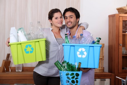 A young couple standing indoors in a home setting, each holding a plastic recycling bin filled with empty glass bottles. The woman on the left holds a bright green bin with a blue recycling symbol, containing clear and green glass bottles of various shapes. The man on the right holds a light blue bin with a similar recycling symbol, also filled with glass bottles, alongside a smaller blue container with punched holes, containing additional green bottles. They are smiling and appear to be preparing for recycling, standing in front of a wooden sideboard with a beige curtain or wall behind them. The environment suggests a kitchen or utility room, with warm lighting and wooden furnishings, supporting a context of household waste management consistent with rubbish and recycling services in Plumstead or nearby areas.