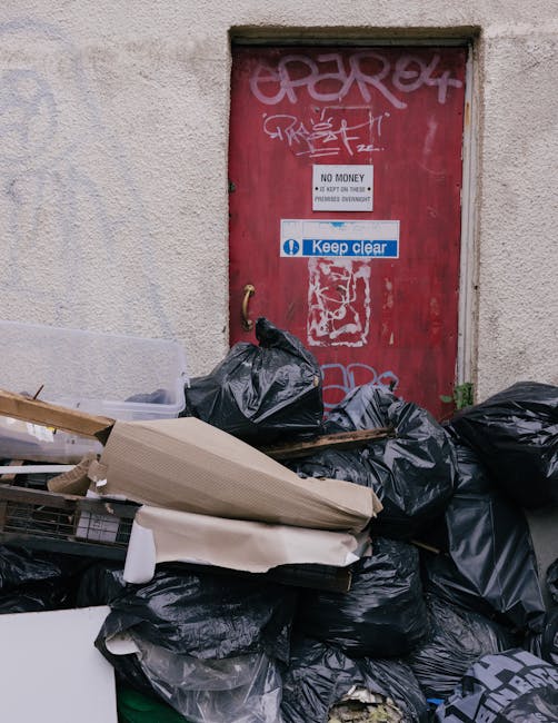 A person wearing a yellow and grey plaid shirt, grey trousers, and light green gloves is holding open a large black rubbish bag with the top rolled over, preparing for waste disposal. The scene is set