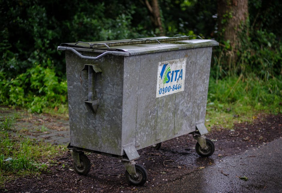 A large, rectangular metal rubbish bin with a textured, weathered grey surface and a closed lid is situated on a patch of wet, dark asphalt pavement outdoors. The bin has four small castor wheels bene
