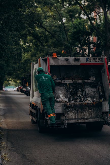 A large, rectangular metal rubbish bin with a textured, weathered grey surface and a closed lid is situated on a patch of wet, dark asphalt pavement outdoors. The bin has four small castor wheels bene